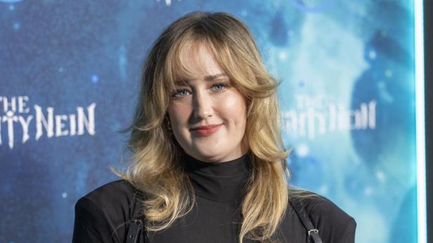 Ashley Johnson lächelt vor einem blauen Hintergrund mit dem Schriftzug „The Mighty Nein“ in die Kamera.