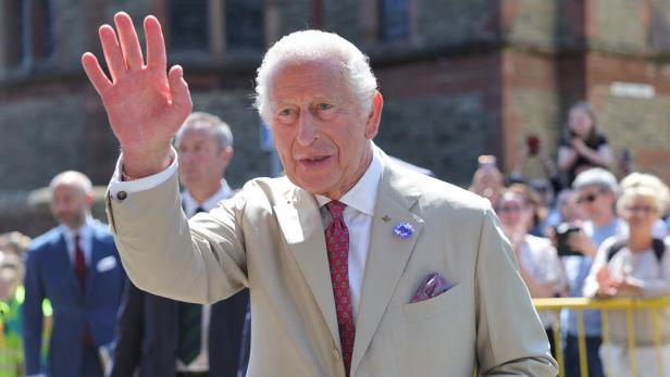 König Charles III. winkt in einem beigen Anzug und mit einer roten Krawatte einer Menschenmenge zu.