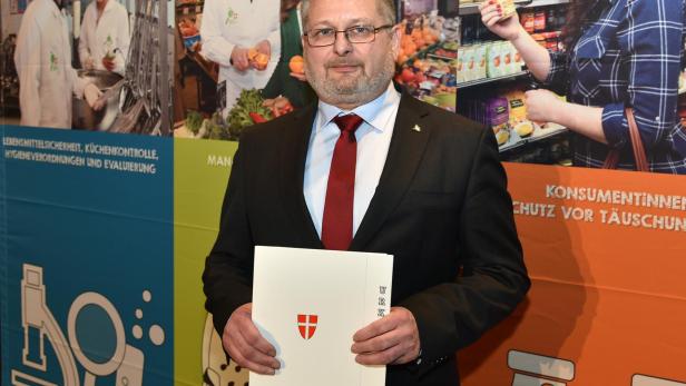 Andreas Kutheil steht vor einer Marktamt-Wandgrafik und hält Unterlagen mit Wiener Wappen in den Händen.