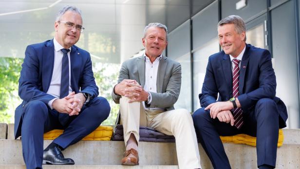 Drei Männer in Business-Kleidung sitzen nebeneinander auf einer Betontreppe vor einem modernen Gebäude.