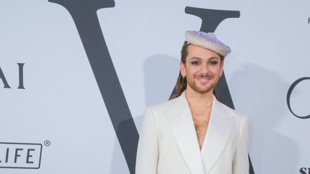 Riccardo Simonetti trägt einen weißen Anzug und eine verzierte Kopfbedeckung vor einer Wand der Vienna Awards for Fashion & Lifestyle.