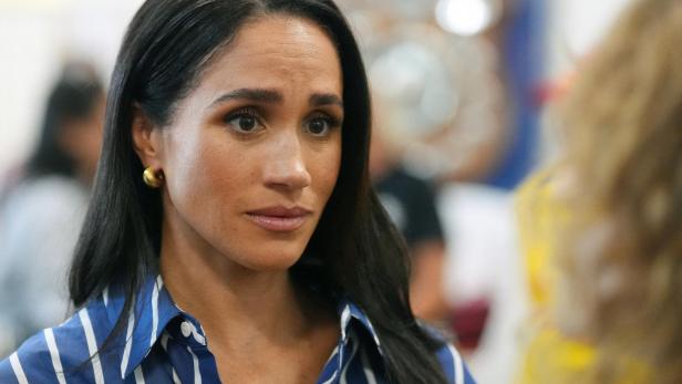 Meghan Markle trägt ein blau-weiß gestreiftes Hemd und blickt mit ernstem Ausdruck zur Seite.