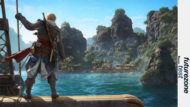 Edward Kenway steht auf dem Schiffsdeck und blickt auf eine tropische Inselbucht mit Felsen, Palmen und einem Dorf.
