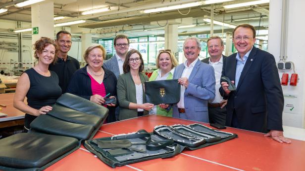 Acht Personen in einer Werkstatt zeigen und halten gepolsterte schwarze Sitzteile auf einem roten Tisch.