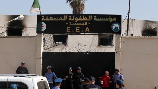 Vor einem Gebäude mit arabischem Schild stehen mehrere Polizisten, ein Mann in Rot und Fahrzeuge; über dem Tor weht eine algerische Flagge.
