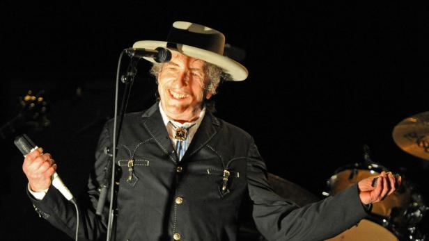 Bob Dylan lächelt auf einer Bühne vor einem Schlagzeug, trägt einen Hut und eine schwarze Jacke mit Bolo-Krawatte und hält ein Mikrofon in der Hand.