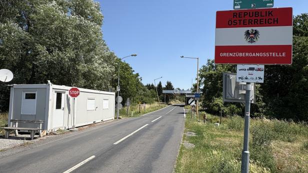 Grenzübergang nach Österreich mit Straßenschild, Kontrollhäuschen und leerer Landstraße unter klarem Himmel.