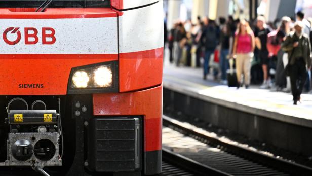Die Front eines rot-weißen ÖBB-Zuges von Siemens steht an einem Bahnsteig, auf dem unscharf erkennbare Menschen warten.