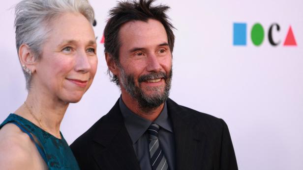 Alexandra Grant und Keanu Reeves lächeln gemeinsam vor einem hellen Hintergrund mit dem MOCA-Logo.