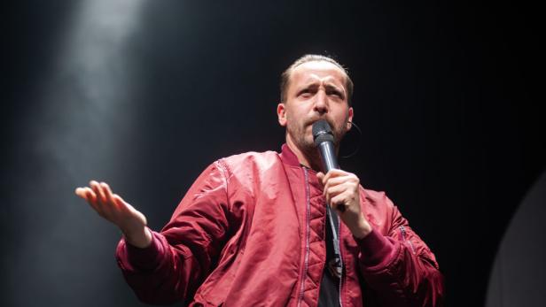 Danger Dan steht mit Mikrofon auf einer dunklen Bühne und gestikuliert während eines Auftritts.