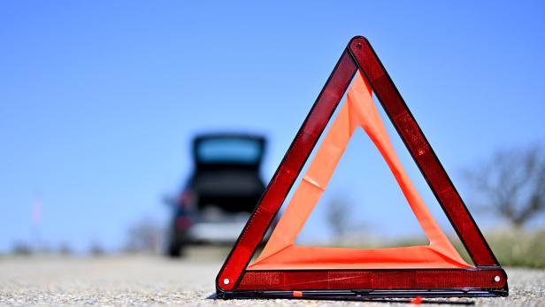 Ein rotes Warndreieck steht auf einer Straße vor einem Auto mit geöffnetem Kofferraum.