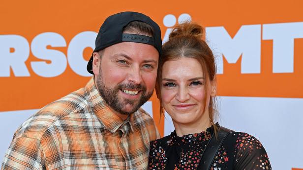 Felix von Jascheroff lächelt neben seiner Ehefrau Alexandra-Sophie Winterberg vor einer orangen GZSZ-Medienwand.