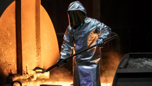 Schweißer in Hitzeschutzkleidung arbeitet mit einer langen Stange vor glühendem Metall und Feuer.