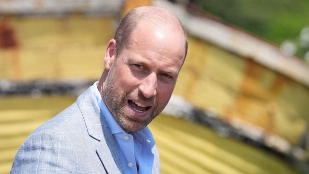 Prinz William trägt ein hellblaues Hemd und ein graues Sakko vor einem unscharfen, gelblichen Hintergrund.