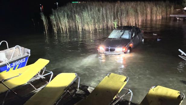 Nachts steckt ein dunkles Auto im flachen Wasser am Ufer, davor liegen gelbe Liegen auf einem Steg.