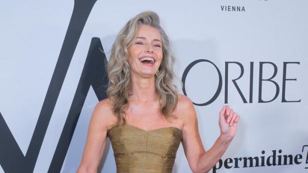 Paulina Porizkova mit grauem Haar lacht und trägt ein trägerloses, goldfarbenes Kleid mit Stufenrock vor einer Sponsorenwand.
