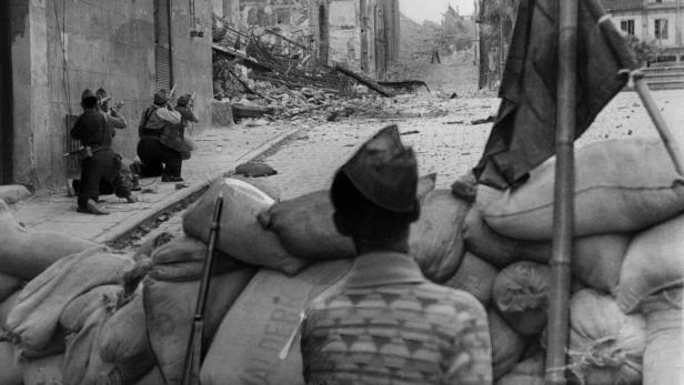 Bewaffnete Soldaten hinter Sandsäcken beobachten eine zerstörte Straße mit Trümmern und Ruinen.