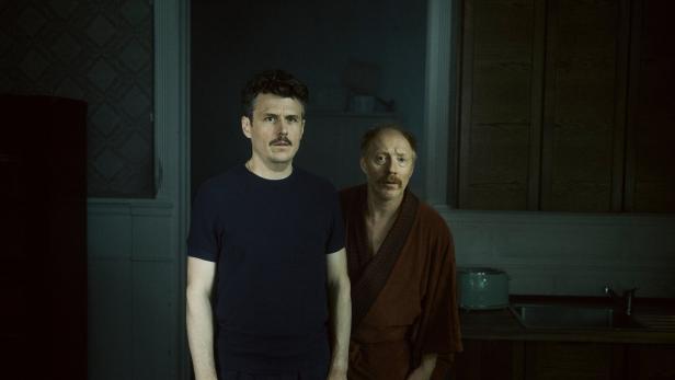 Manuel Rubey und Simon Schwarz stehen in einer dunklen Küche und blicken ernst in die Kamera.