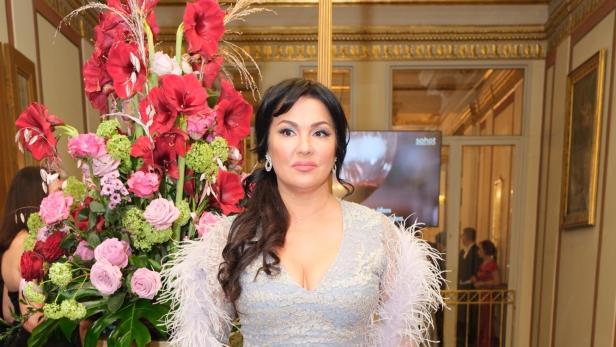 Anna Netrebko trägt ein hellblaues, mit Federn verziertes Abendkleid neben einem großen Blumenstrauß vor einer Treppe.