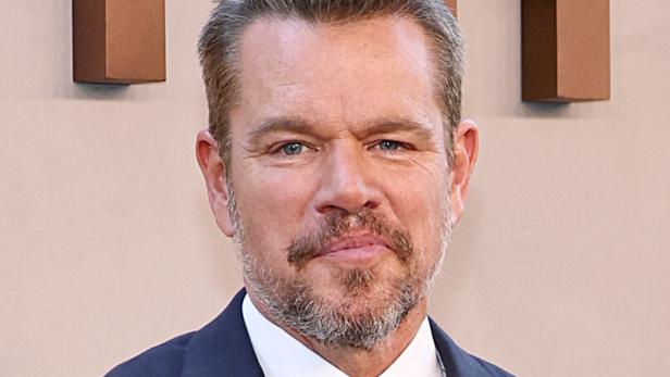 Matt Damon trägt einen dunkelblauen Anzug mit weißem Hemd und einer gemusterten Krawatte vor einem hellen Hintergrund.