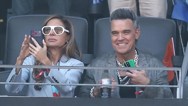 Ayda Field und Robbie Williams sitzen lächelnd auf der Tribüne, während Ayda ein Foto mit ihrem Smartphone aufnimmt.