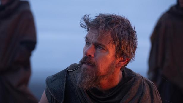Matt Damon mit Bart und in historischer Kleidung blickt vor einem bläulichen Hintergrund nach oben, während sein Gesicht von warmem Licht beleuchtet wird.