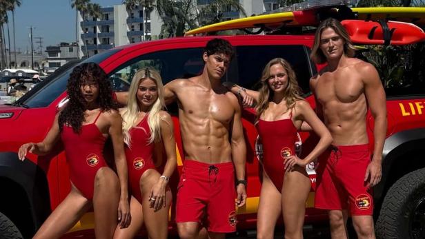 Fünf Rettungsschwimmer in roten Badeanzügen und Shorts posieren vor einem roten Lifeguard-Pick-up mit Surfbrettern auf dem Dach.