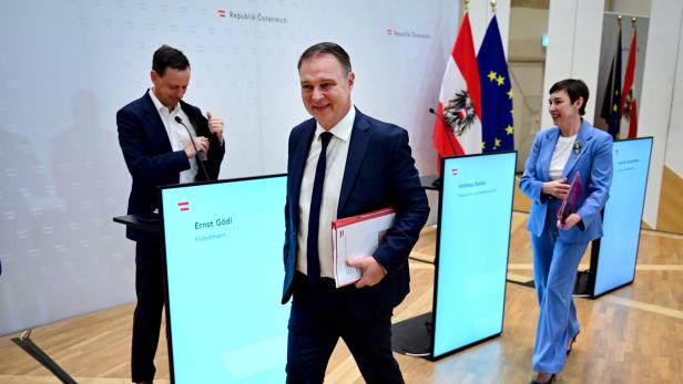 Andreas Babler geht mit einer Mappe durch einen Raum, in dem Ernst Gödl und Henrike Brandstötter  vor Infoständern stehen.