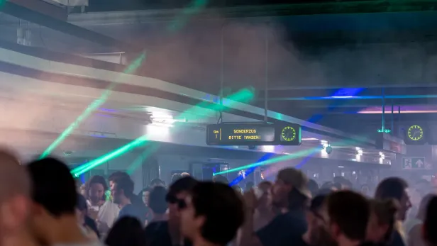 In einer U-Bahn Station sind mehrere Leute auf einem Techno Event, in der Luft sind Nebel und Lichter.
