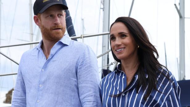 Prinz Harry mit einer Invictus-Games-Kappe und Meghan Markle stehen vor Segelbooten.