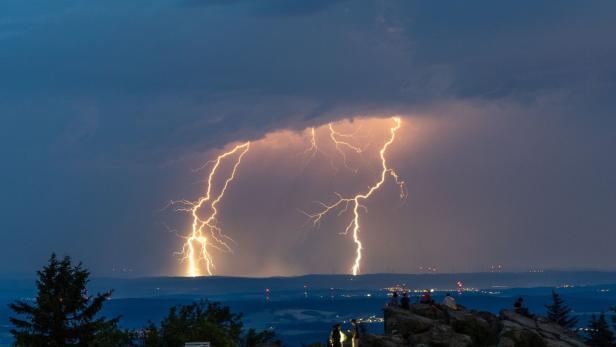 Gewitter über der Landschaft, zwei helle Blitze leuchten am Horizont, darunter stehen Menschen auf Felsen.
