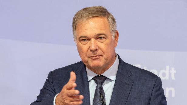 Ein Mann im Anzug spricht mit ausgestreckter Hand vor hellviolettem Hintergrund.