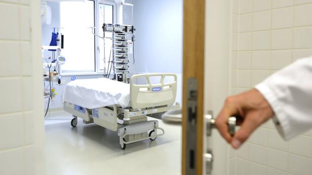 Operationssaal mit leerem Krankenhausbett und medizinischen Geräten, eine Hand öffnet die Tür.