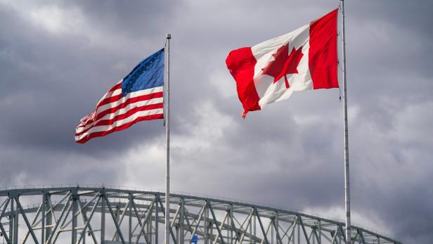 US- und kanadische Flagge wehen vor einer Stahlbrücke unter grauem Himmel.