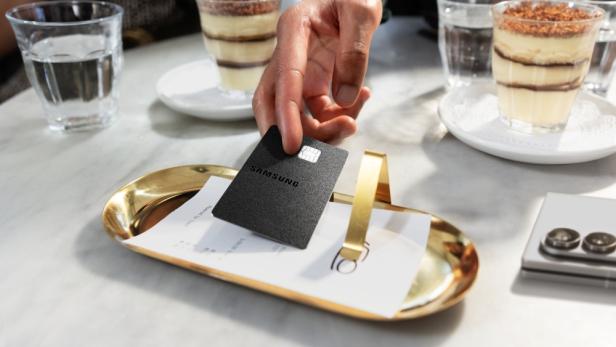 Schwarze Samsung Kreditkarte liegt auf einem goldenen Tablett mit der Rechnung, im Hintergrund stehen Gläser mit Dessert und Wasser