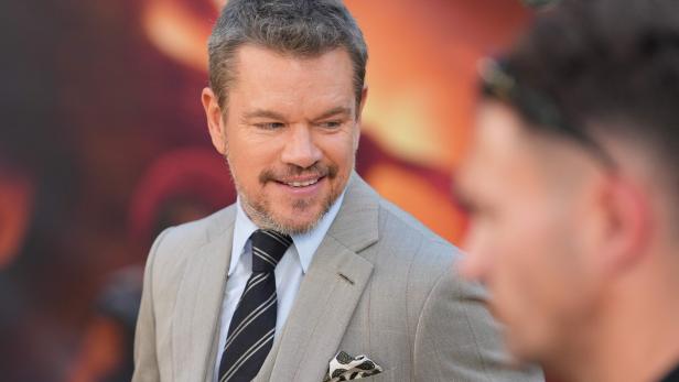 Matt Damon lächelt in einem hellen Anzug mit Krawatte vor einem unscharfen, orangefarbenen Hintergrund.