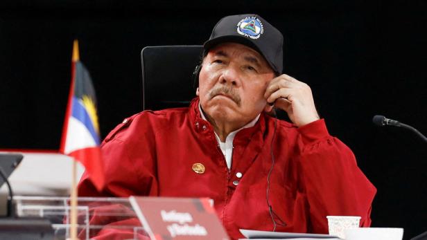 Daniel Ortega trägt eine schwarze Kappe mit dem Wappen Nicaraguas und eine rote Jacke, während er an einem Tisch mit einer Flagge sitzt.