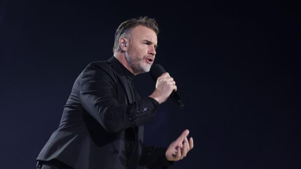 Gary Barlow singt vor dunklem Hintergrund in ein Mikrofon.