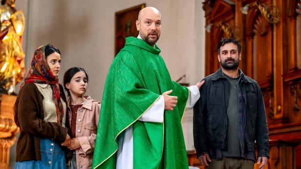 Ein Priester in grünem Messgewand steht in einer Kirche neben einer Familie und legt einem Mann die Hand auf die Schulter.
