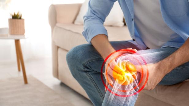 Mann sitzt auf dem Sofa und hält schmerzhaft sein Knie, markiert mit rotem Schmerzsymbol.