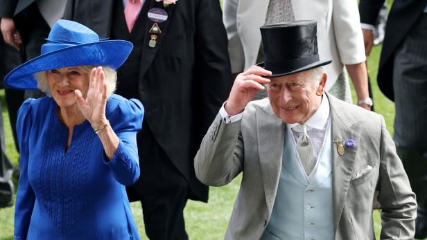 König Charles III. und Königin Camilla winken bei einem öffentlichen Auftritt, wobei Charles seinen Zylinder leicht anhebt.