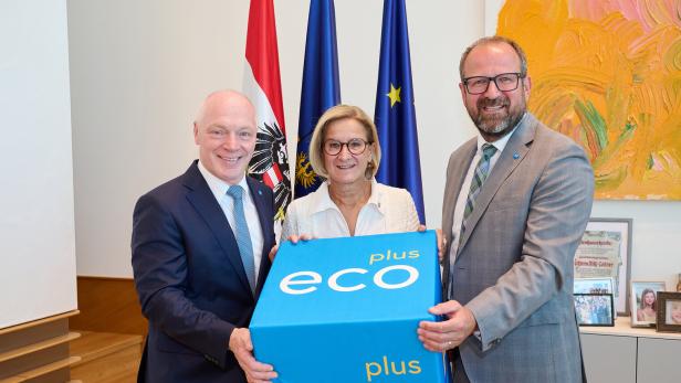 Drei Personen halten gemeinsam einen blauen „eco plus“-Würfel vor Fahnen in einem Büro.