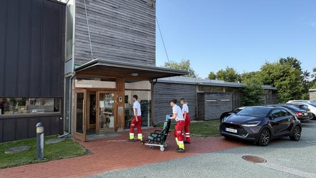 Drei Rettungskräfte bringen eine Trage vor ein modernes Gebäude.