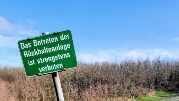 Grünes Verbotsschild an einem Rückhaltebecken vor hügeliger Wiese und blauem Himmel.