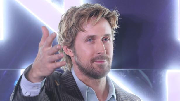 Ryan Gosling trägt ein gestreiftes Sakko und ein helles Hemd und streckt seine Hand vor einem leuchtenden Hintergrund aus.