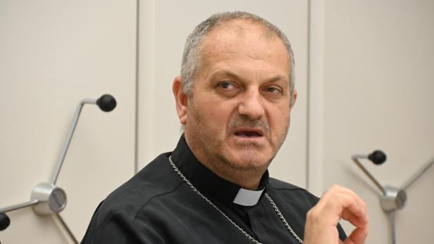 Der syrische Erzbischof Jacques Mourad sitzt in schwarzem Priestergewand vor einer weißen Wand.