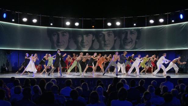 Ballett-Tänzerinnen und Tänzer bewegen sich synchron auf einer Bühne, im Hintergrund sind die Beatles projeziert.