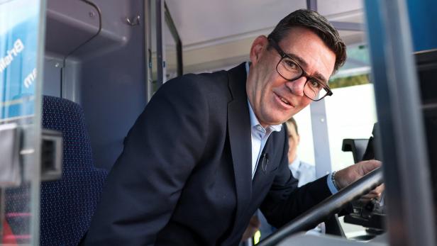 Mann im Anzug mit Brille sitzt am Steuer eines Busses und lächelt in die Kamera.