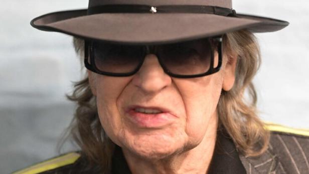 Udo Lindenberg trägt einen grauen Hut, eine Sonnenbrille, ein schwarzes Hemd mit Krawatte und ein gestreiftes Sakko.
