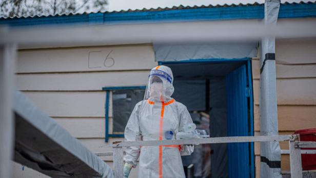 Person in Schutzanzug und Gesichtsschild steht vor einem Gebäude mit blauem Eingang.
